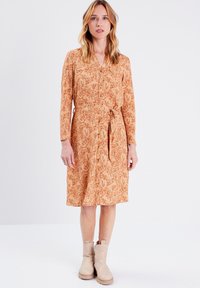 Langärmliges, knopfbares Kleid in Orange mit floralem Muster, an der Taille mit einem Gürtel gerafft. Ergänzt durch beigen Knöchelstiefel.