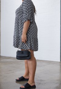 Kvinde iført mønstret knælang kjole og sorte slidset sandaler, mens hun holder en lille sort quiltet håndtaske med kædetræk, foran en murstensvæg.