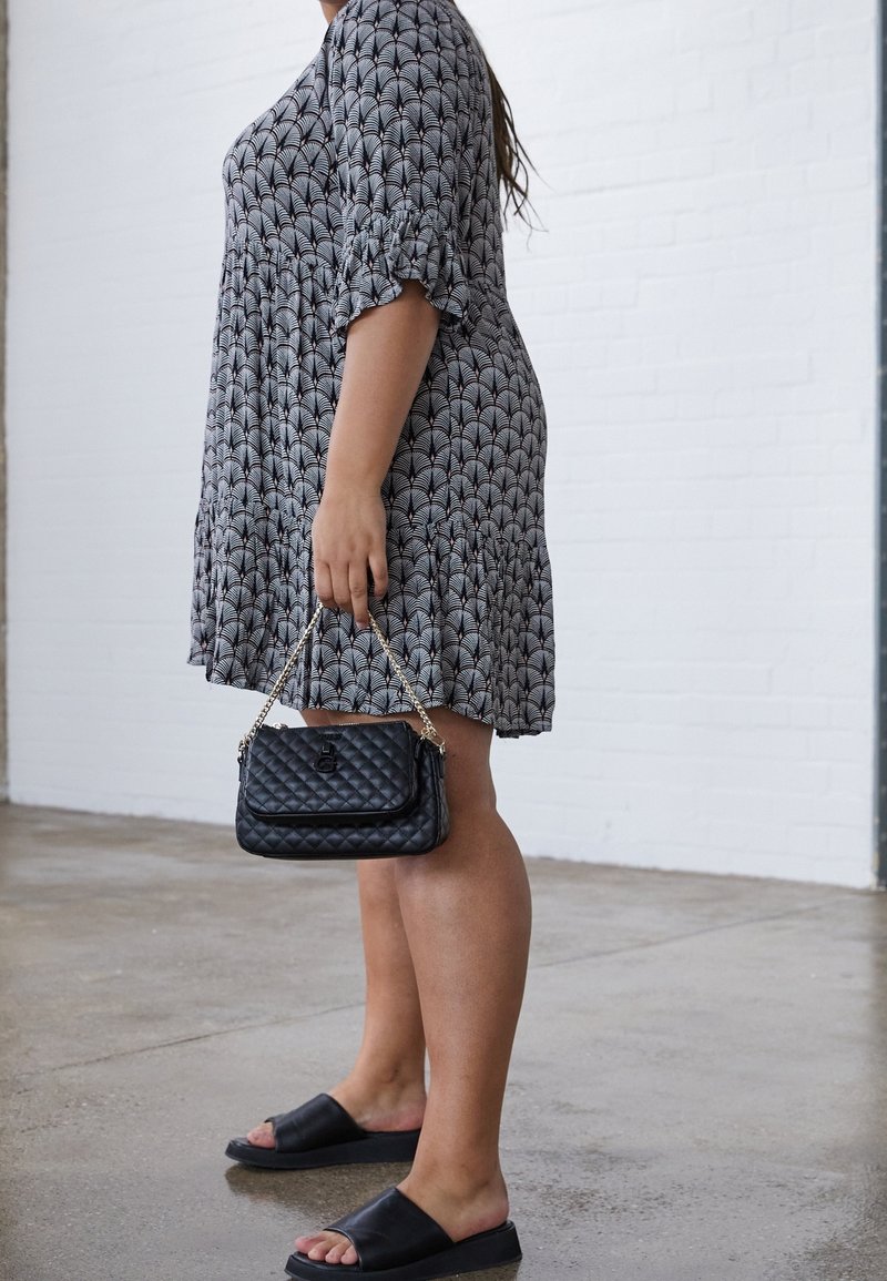 Kvinde iført mønstret knælang kjole og sorte slidset sandaler, mens hun holder en lille sort quiltet håndtaske med kædetræk, foran en murstensvæg.