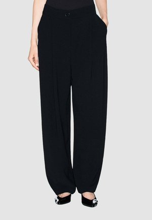 Persona che indossa pantaloni neri larghi con tasche laterali e scarpe basse nere e bianche con punta, in piedi davanti a uno sfondo chiaro e uniforme.