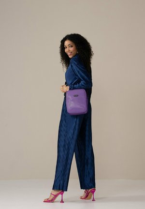 Paars accessoire-tasje met een gestructureerd oppervlak, met een rechthoekige vorm en zwart logo. Gedorst over de schouder met een marineblauwe outfit.
