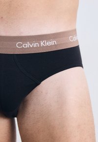 Zwarte katoenen slip met een bruine tailleband waarop "Calvin Klein" in het wit is gedrukt. Glad materiaal en een aansluitend ontwerp dat de silhouet benadrukt.