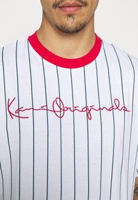 Bílé pruhované tričko s červeným límečkem. Obsahuje modré svislé pruhy a vyšitý nápis "Kani Originals" červenou barvou na přední straně. Měkká bavlněná tkanina.