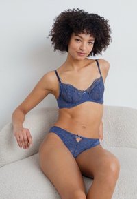 Lace lingerie set in deep blue. The bra features sheer panels, decorative hardware, and thin straps. The matching brief has a small bow detail.