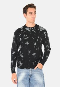 Maglione nero con un motivo astratto grigio testurizzato; scollo rotondo, polsini e orlo a coste, abbinato a jeans azzurri chiari.