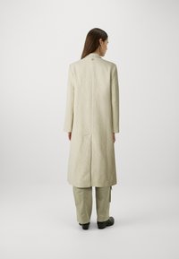 Beige kabát ve vzoru herringbone s dlouhými rukávy a klopovým límcem, s rozparkem na zadní straně a zlatými detaily u krku. Nosený přes zelené kalhoty.