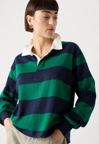 Rugby-stijl top met groene en marineblauwe strepen, voorzien van een witte kraag en een ontspannen pasvorm. Lange mouwen met geribde manchetten. Zachte textuur.