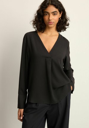 Blouse noire à manches longues avec un col en V, présentant une coupe ample et un léger drapé, fabriquée en tissu doux et léger.