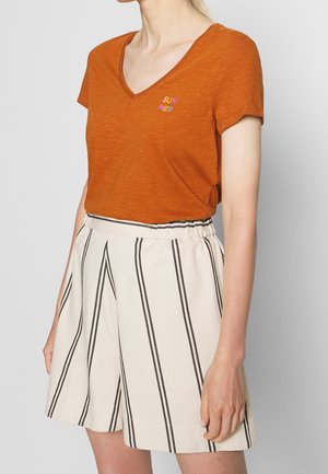 Vrouw draagt een verbrand oranje v-hals T-shirt met de tekst "SUMMER" en een witte, hooggetailleerde short met verticale zwarte strepen.
