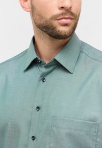 Camicia button-up verde chiaro con un tessuto texture, bottoni scuri a contrasto e una tasca sul petto. Presenta un colletto a motivi blu.