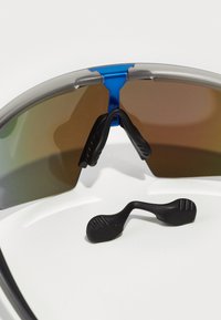 Sportsonnenbrille mit silbernem Rahmen, braun getönten Gläsern und blauer Brücke. Ein abnehmbarer Nasenpolster aus schwarzem Gummi ist enthalten.