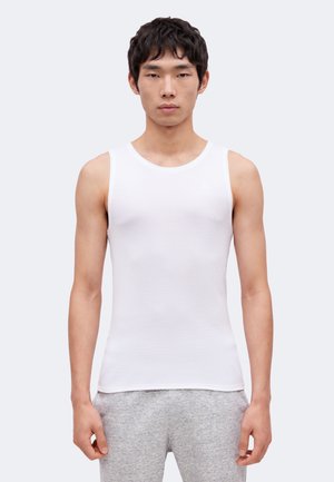 Joven con cabello corto y oscuro que lleva una camiseta blanca sin mangas ajustada y pantalones deportivos gris claro, de pie frente a un fondo sencillo.
