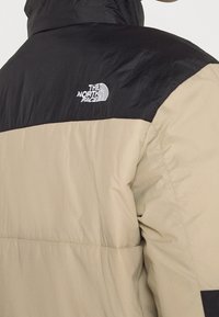 Beige och svart pufferjacka, med quiltad textur, hög krage och logotyp på vänster axel. Lätt och isolerad design.