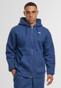 Marineblauwe rits-hoodie gemaakt van zacht materiaal, met een kangoeroezak, ribgebreide manchetten en een klein logo op de linkerkant van de borst.