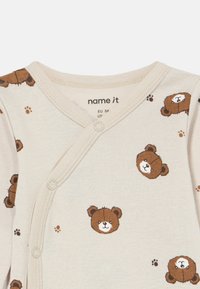 Lys beige babybodie med brune bjørneansiktstrykk, trykknapper og mykt bomullsmateriale. Har en overlappende frontdesign.