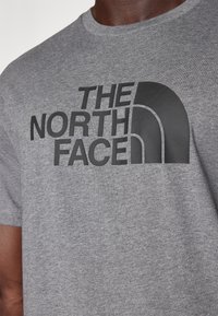 Camiseta de algodón gris con un gran logo negro que presenta el texto "THE NORTH FACE" y un distintivo elemento de diseño curvado. Textura de tela suave.