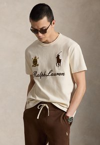 Άνδρας που φοράει μπλούζα Ralph Lauren κρεμ με λογότυπα και καφέ παντελόνι με κορδόνι, γυαλιά ηλίου και ρολόι χειρός, κοιτάζοντας κάτω με τα χέρια στις τσέπες.
