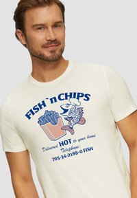 Witte T-shirt met een blauw en oranje grafisch ontwerp van een vis en friet, met de tekst "FISH 'n CHIPS Warm bij je thuis bezorgd" en een telefoonnummer.