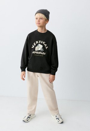 Garçon portant un bonnet gris, un sweat-shirt noir avec le texte "Vertical Adventure" et un graphisme d'escalade, un pantalon beige et des baskets blanches, debout sur un sol blanc.