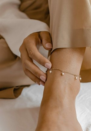 Gold-colored ankle bracelet with small, evenly spaced white pearls, worn on a foot. The skin is tan, with polished nails visible.