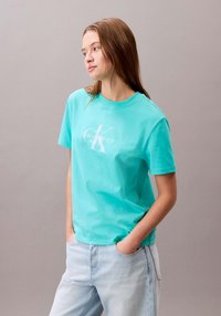 T-shirt in cotone turchese con maniche corte, con il logo Calvin Klein bianco stampato sul petto. Abbinata a jeans azzurri.