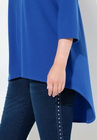 Maglietta blu oversize con orlo arrotondato, abbinata a jeans scuri con dettagli in metallo lungo il lato. Texture liscia.