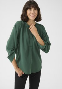 Blusa verde a maniche lunghe con scollatura rotonda, patta frontale e polsini arricciati. Tessuto liscio, design semi-adatto e tessuto leggero.