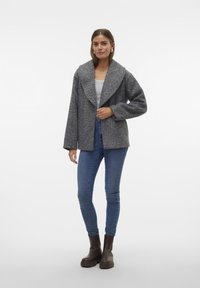Veste en mélange de laine grise avec un col châle et une ouverture sur le devant, portée sur un haut clair, associée à un jean bleu et des bottes à talons brunes.