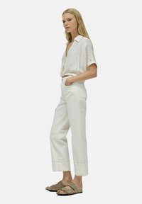 Camisa blanca texturizada metida en pantalones blancos de tiro alto con un dobladillo recortado. La modelo lleva sandalias marrones y está de pie con una mano en el bolsillo.