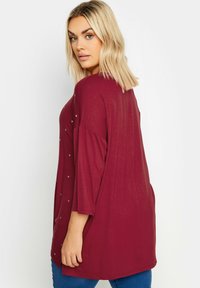 Burgundy långärmad topp med avslappnad passform, prydd med stjärnformade detaljer på vänster sida. Mjuk material och medellånga ärmar.