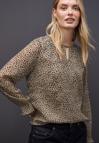 Vrouw met blond haar, gekleed in een beige blouse met lange mouwen en luipaardprint, en donkere spijkerbroek, tegen een donkergrijze achtergrond.