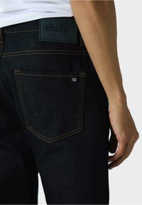 Temno modri traper hlače s ožjim krojem, opremljene z oranžnim šivom, logotipom na pasu in minimalističnimi zadnjimi žepi.