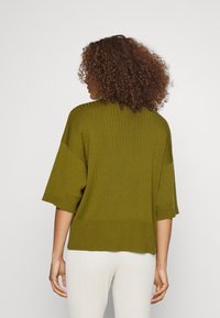 Olivově zelený pletený svetr s krátkým rukávem, žebrovanou strukturou a volným střihem, se rovnými lemy a bočními rozparky.