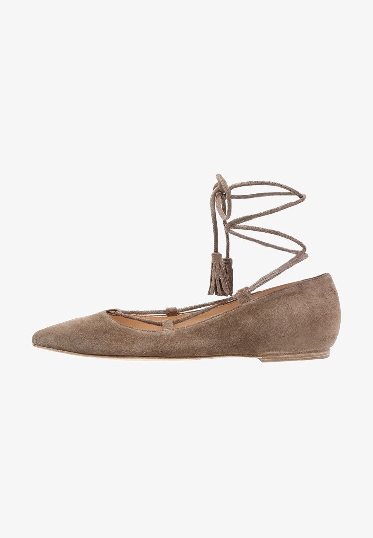 Chaussure plate en daim taupe à bout pointu avec des lanières enroulées autour de la cheville se terminant par des pompons, présentée sur un fond blanc.