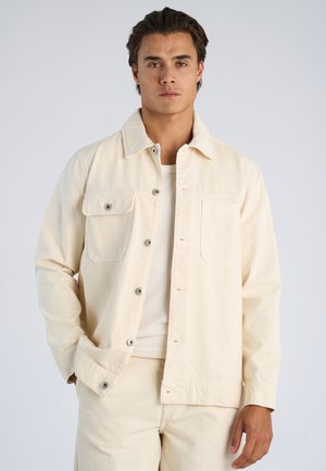 Młody mężczyzna w kremowej kurtce na guziki, nałożonej na dopasowaną jasną koszulę i spodnie, stoi na tle jednolitego jasnoszarego tła.