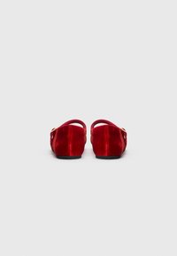 Un par de zapatos planos de terciopelo rojo con tira trasera y hebilla dorada, mostrados desde la parte trasera sobre un fondo gris claro y liso.