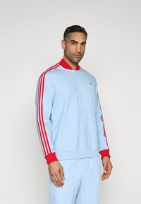 Light blue sweatshirt with a red ribbed collar, red shoulder stripes, and subtle logo detail on the chest. Soft, textured fabric.