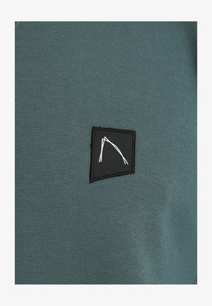 Dunkelgrüner Sweatshirtstoff mit glatter Textur, der einen kleinen schwarzen Aufnäher mit einem weißen geometrischen Design aufweist.