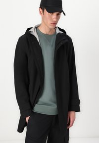 Zwarte, waterdichte jas met een capuchon, voorzien van een gestreepte binnenkant. Draagt over een lichtgroene sweater, met zwarte broek en een pet.