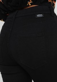 Primer plano de una persona que lleva puestos unos pantalones vaqueros negros de cintura alta con bolsillos traseros y una blusa de estampado floral metida por dentro.