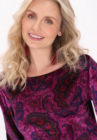 Lila Paisley-musterte Bluse mit weitem Ausschnitt, weicher Textur und lebhaften floralen Akzenten. Hat lange, wellige Haare und hängende Ohrringe.