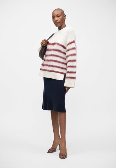 Wit-rood gestreepte trui, pluizige textuur, gecombineerd met een marineblauwe klokrok en bruine hoge hakken. Model houdt een bruine tas vast.