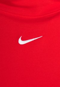 Röd sporttopp med en slät, ribbad textur. Har en vit Nike-logotyp broderad fram. Rund halsringning.