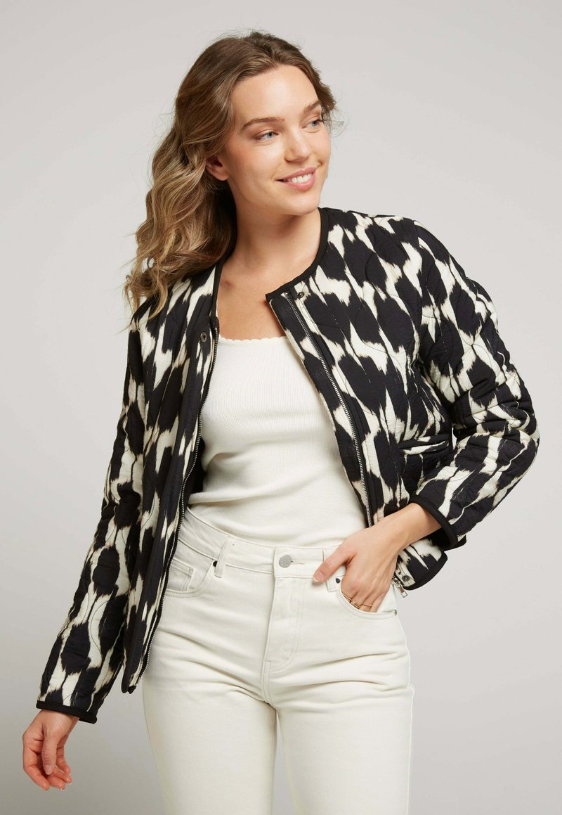 Veste bomber matelassée dans un motif ikat noir et blanc, avec un col rond et une fermeture éclair. Portée sur un haut ajusté blanc et un jean clair.