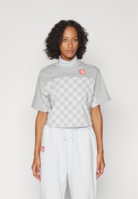 Tricou scurt gri cu un model de tablă de șah în față, având un logo Nike discret și o pată de accent roșie, purtat cu pantaloni de trening gri.