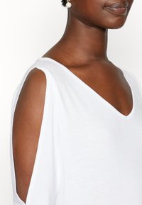 Witte V-hals top met open schouderontwerp, zacht textuur en een relaxed fit, benadrukt door minimale stikseldetails.