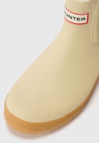 Botte de cheville en caoutchouc beige avec une semelle couleur tan, ornée d'une étiquette rouge et blanche affichant "HUNTER" près du haut. Texture lisse, bout arrondi.