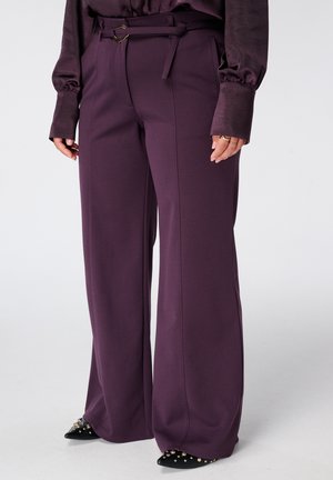 Paarse broek met wijde pijpen en bijpassende riem, gedragen met een donkerpaarse longsleeve en zwarte gespijkerde puntige schoenen.