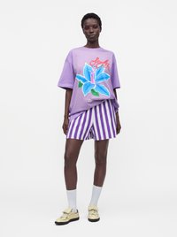 Γυναίκα που φοράει μοβ oversized μπλούζα με μεγάλο μπλε λουλουδένιο σχέδιο, μοβ και λευκή ριγέ σορτς, λευκές κάλτσες και μπεζ αθλητικά παπούτσια.