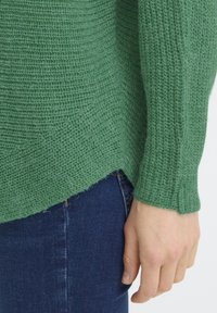 Pull vert en maille avec un tissage texturé et un ourlet légèrement courbé. Associé à un jean en denim bleu. Gros plan sur la manche et la main.